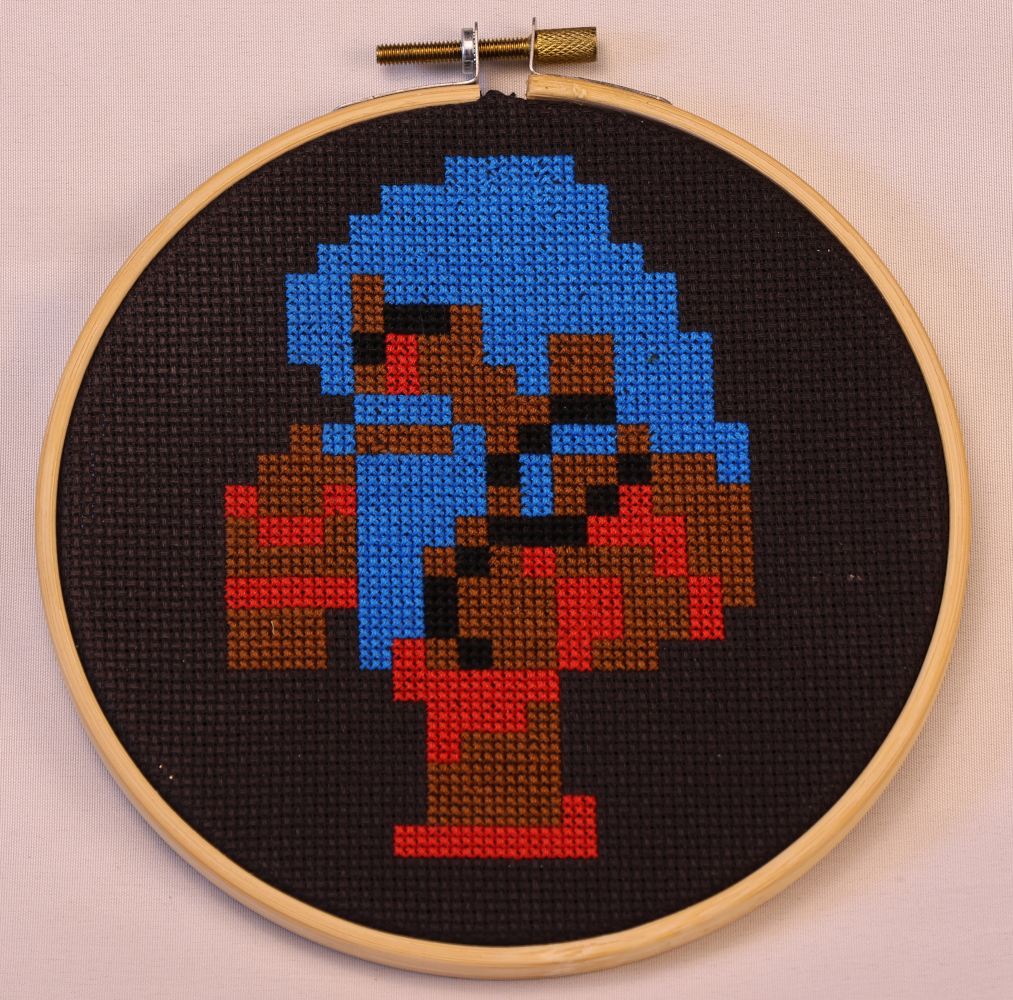 Photograph of a cross stitch on black aida. A pixel-rendered barbarian in the FF1 style with scraggly blue hair, a red eye, wearing a red loin cloth and sandals, and brown skin.
